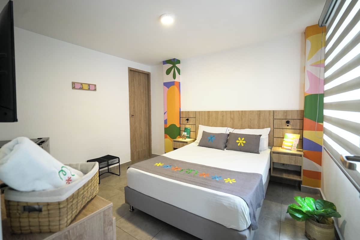 A hotel room with a bed, bedside table, nightstand, TV, and a window with blinds.