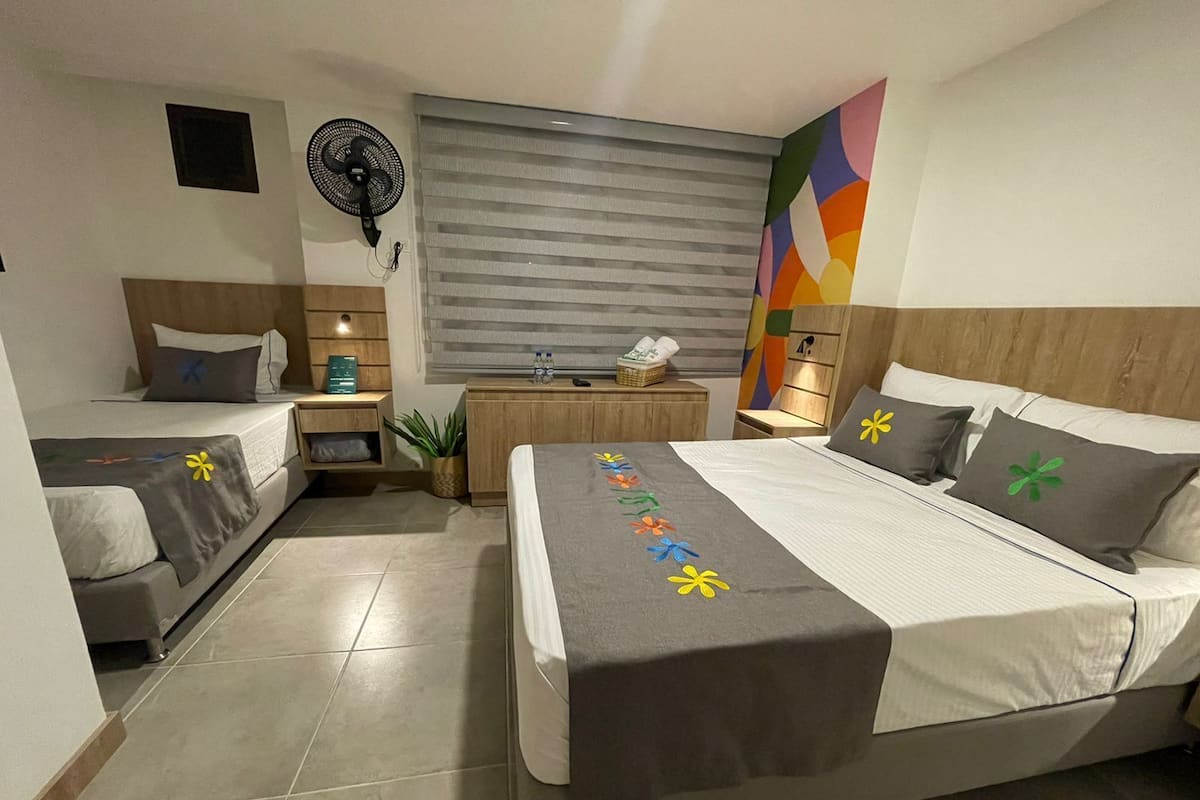 A hotel room with two beds, a desk, a chair, a fan, and a colorful abstract painting on the wall.