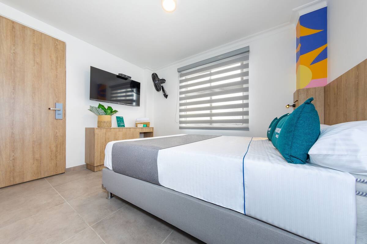 A modern bedroom with a bed, a TV, a window with blinds, a wooden wardrobe, and a small table with a plant.