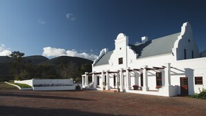 A white colonial-style building with a grey roof, surrounded by a paved area and a white wall with greenery.