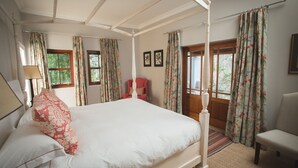 A four-poster bed with a canopy, a red chair, and a white armchair in a room with floral curtains and framed pictures on the walls.