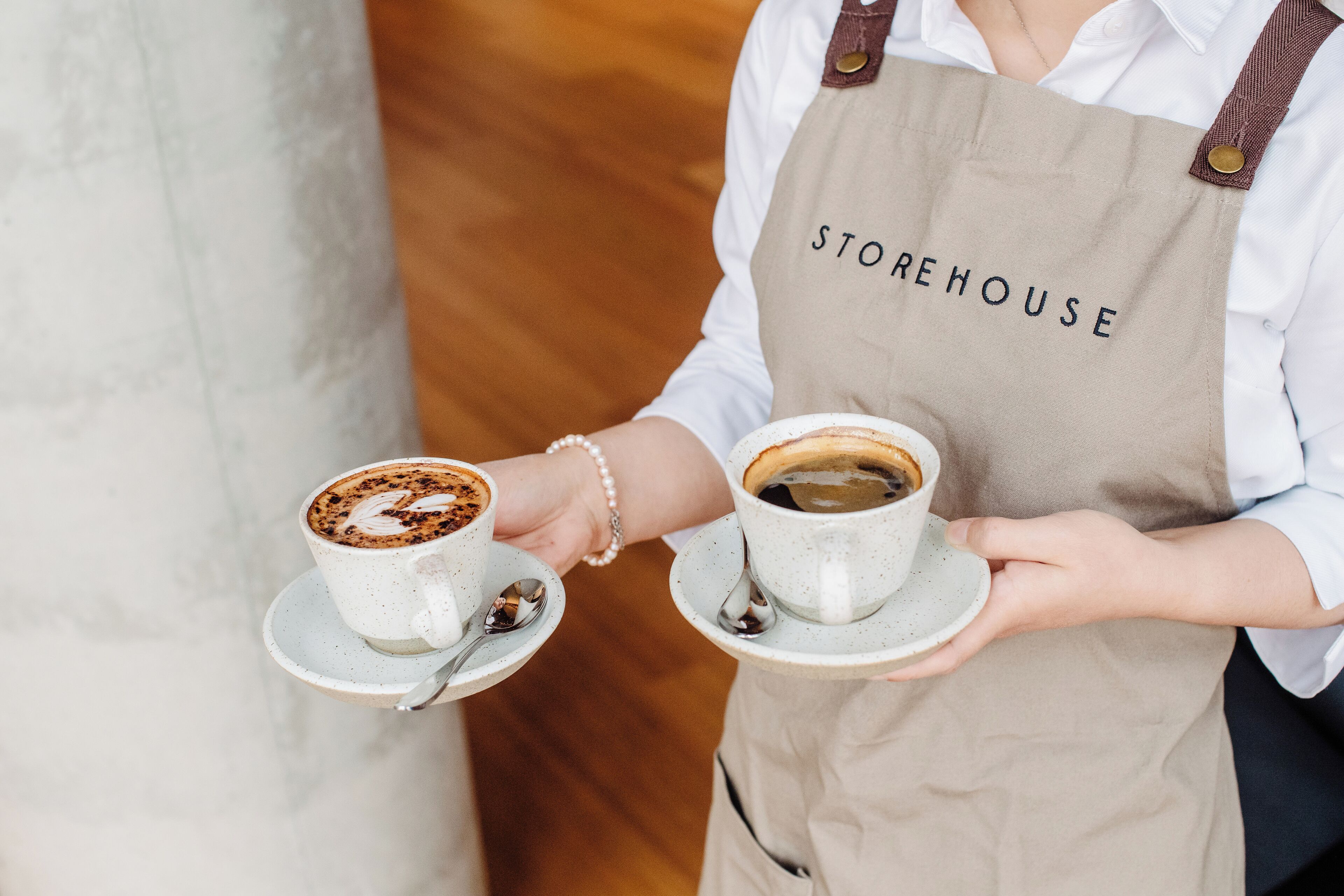 A person holding two cups of coffee, one with latte art, wearing a beige apron with "STOREHOUSE" written on it.