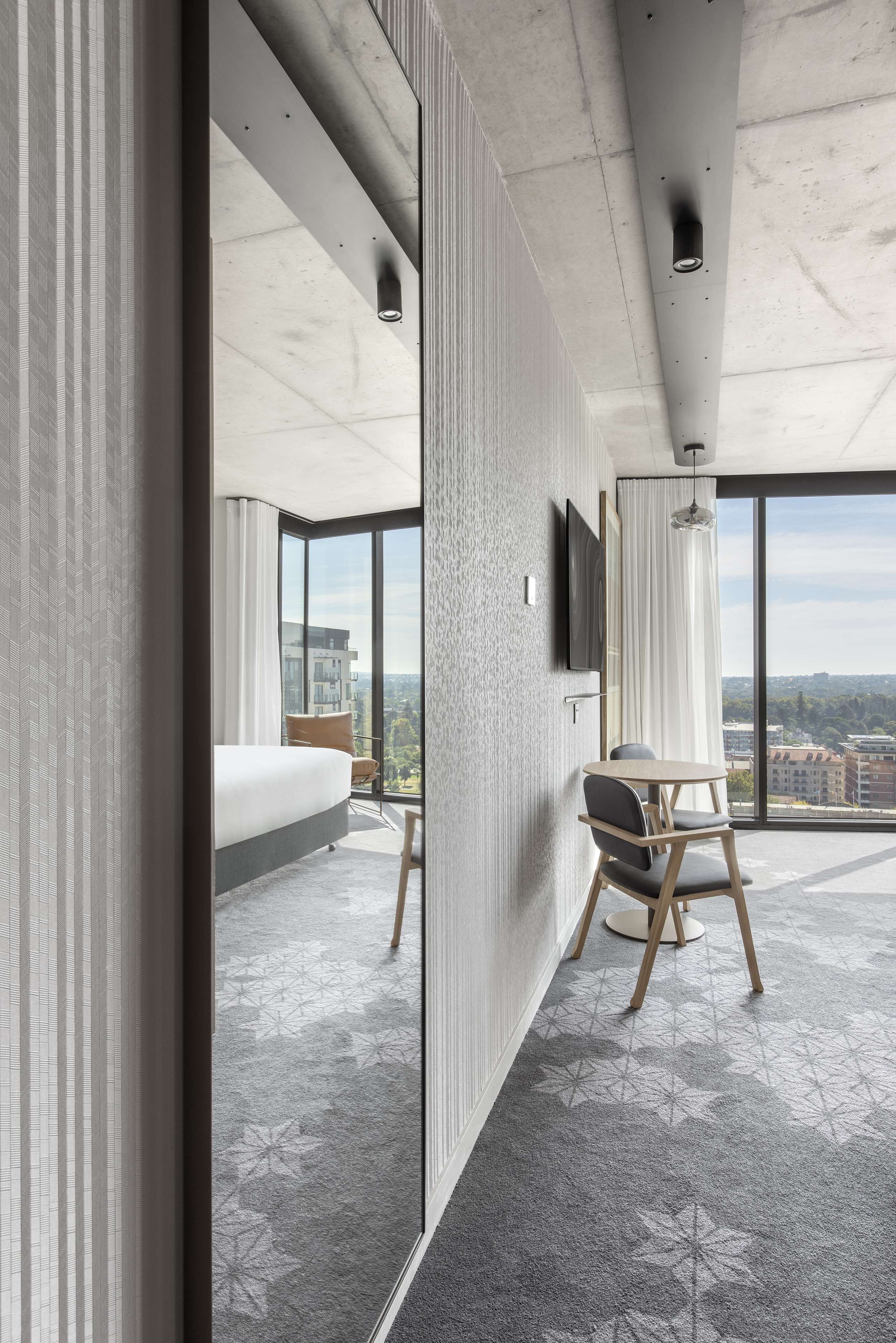 A modern hotel room with a large window, a bed, a chair, and a TV mounted on the wall.