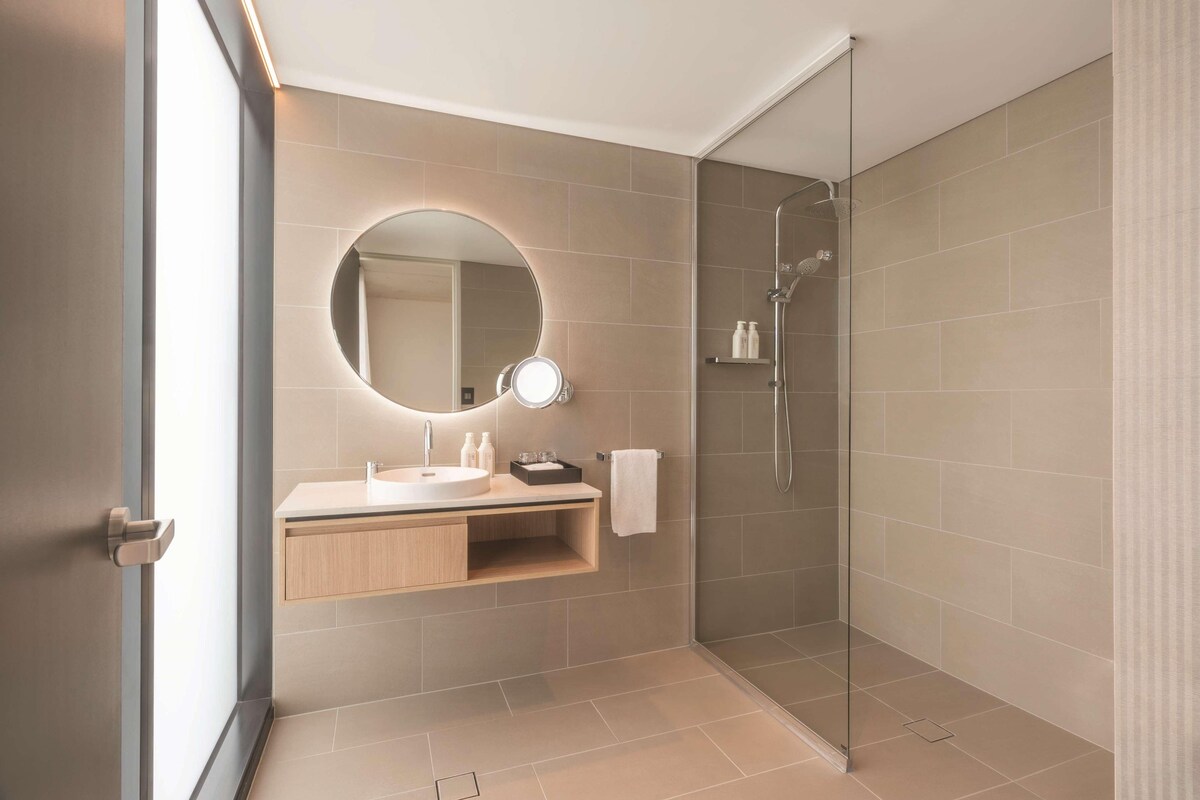 Un baño moderno con ducha de vidrio, un espejo redondo con luz y un tocador de madera.