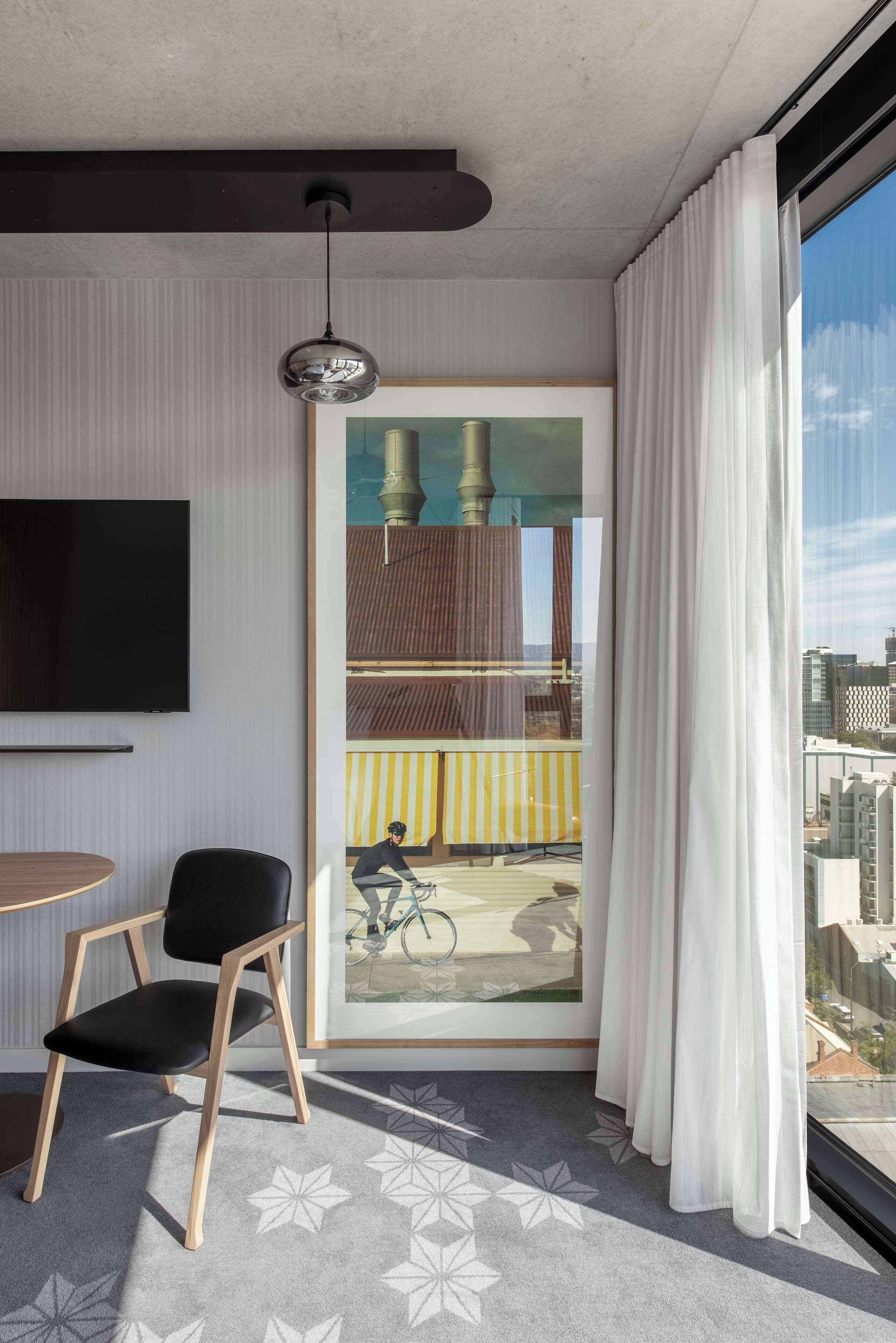 A modern hotel room with a large window, a mounted television, a framed cityscape painting, a black chair, and a patterned carpet.