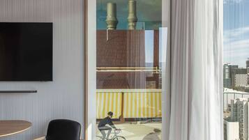 A modern hotel room with a large window, a mounted television, a framed cityscape painting, a black chair, and a patterned carpet.