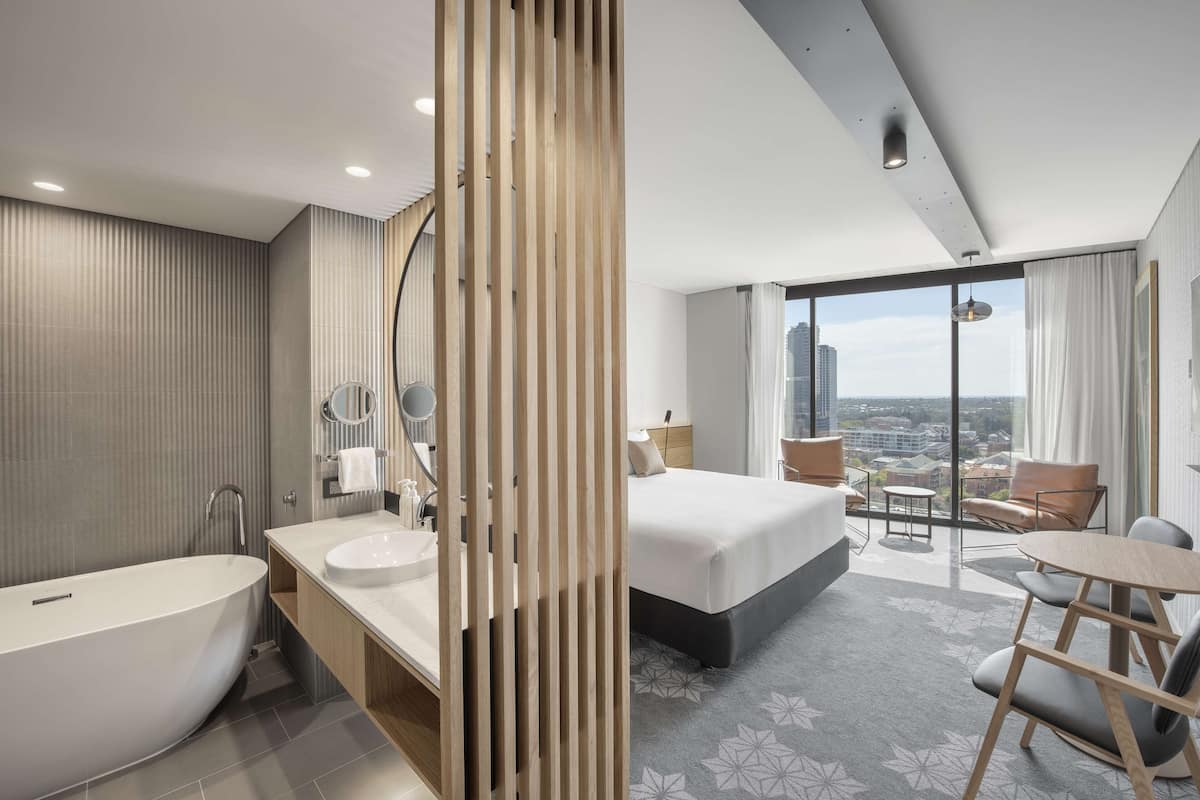 Una habitación de hotel moderna con una cama grande, una bañera, un tocador con espejo y un balcón con vistas a la ciudad.