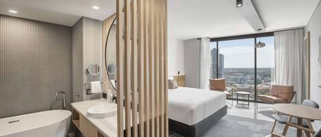 A modern hotel room with a large bed, a bathtub, a vanity with a mirror, and a balcony with a city view.