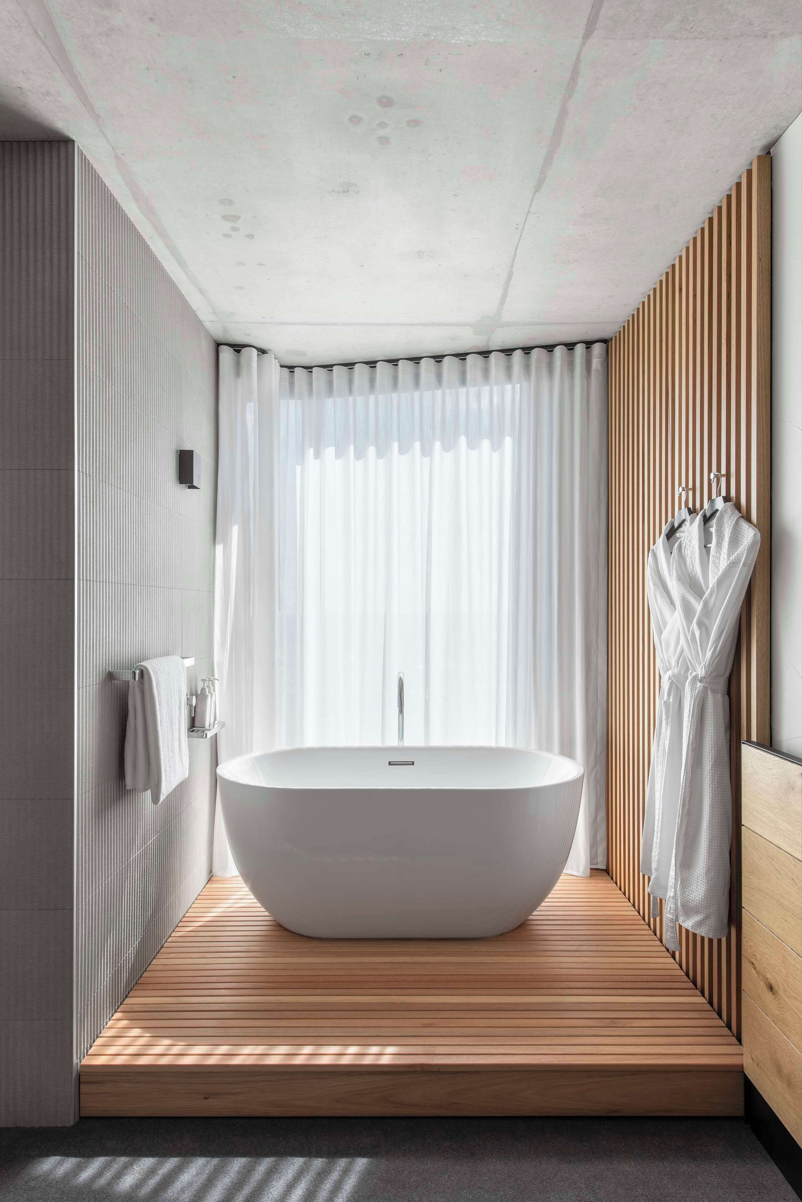 A modern bathroom with a freestanding bathtub, wooden flooring, and a large window with sheer curtains.