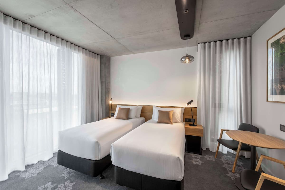 Habitación de hotel con dos camas, un escritorio, una silla y un ventanal con cortinas translúcidas.