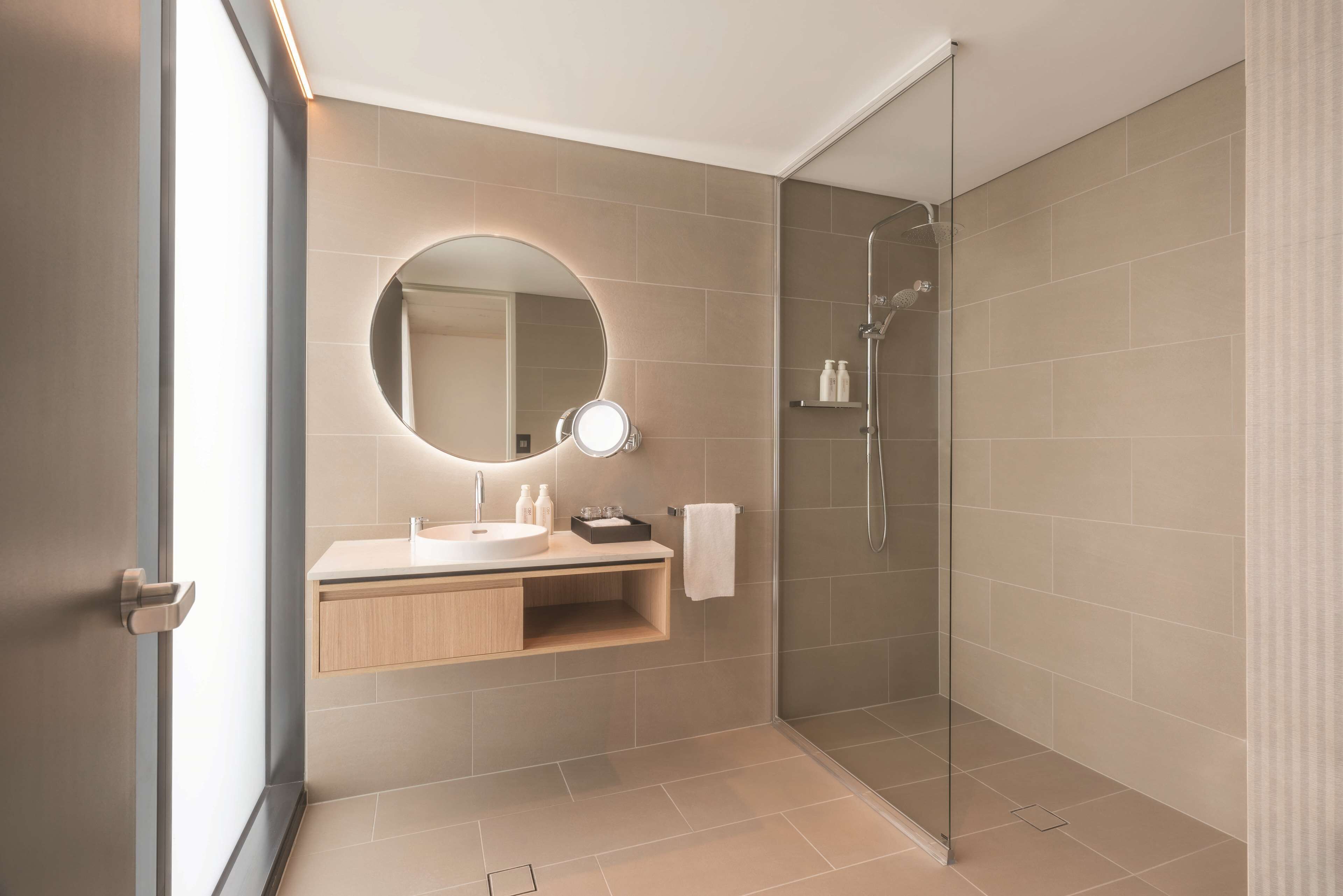 Une salle de bain moderne avec une douche à l’italienne en verre, un grand miroir rond avec éclairage intégré et un meuble-lavabo en bois.