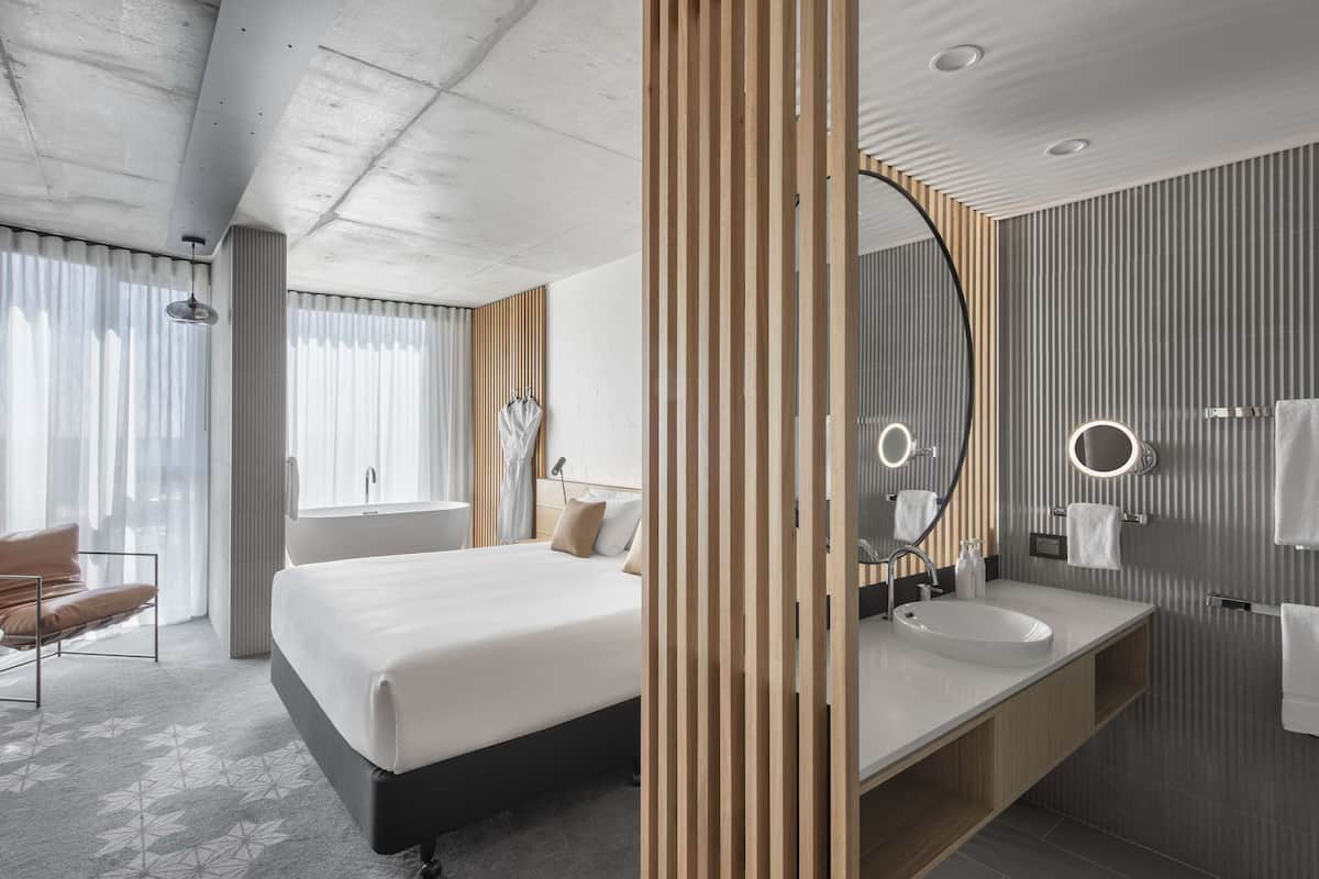Una habitación de hotel moderna con una cama grande, una separadora de madera, un espejo redondo, un lavabo blanco y una ventana con cortinas.