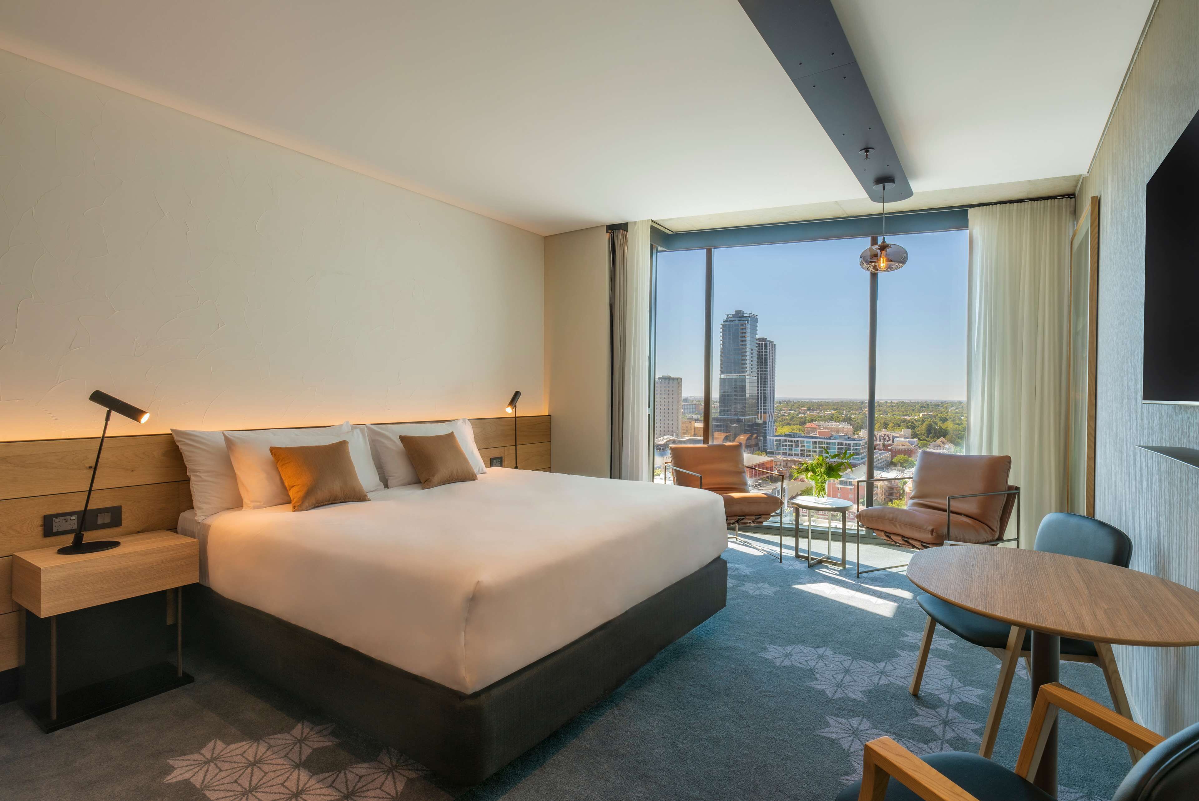 A modern hotel room with a large bed, a desk, a chair, a small table, and a view of the city through large windows.