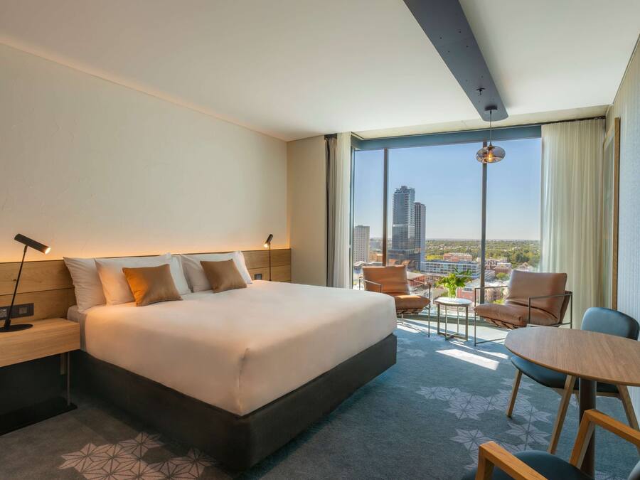 A modern hotel room with a large bed, a desk, a chair, a small table, and a view of the city through large windows.