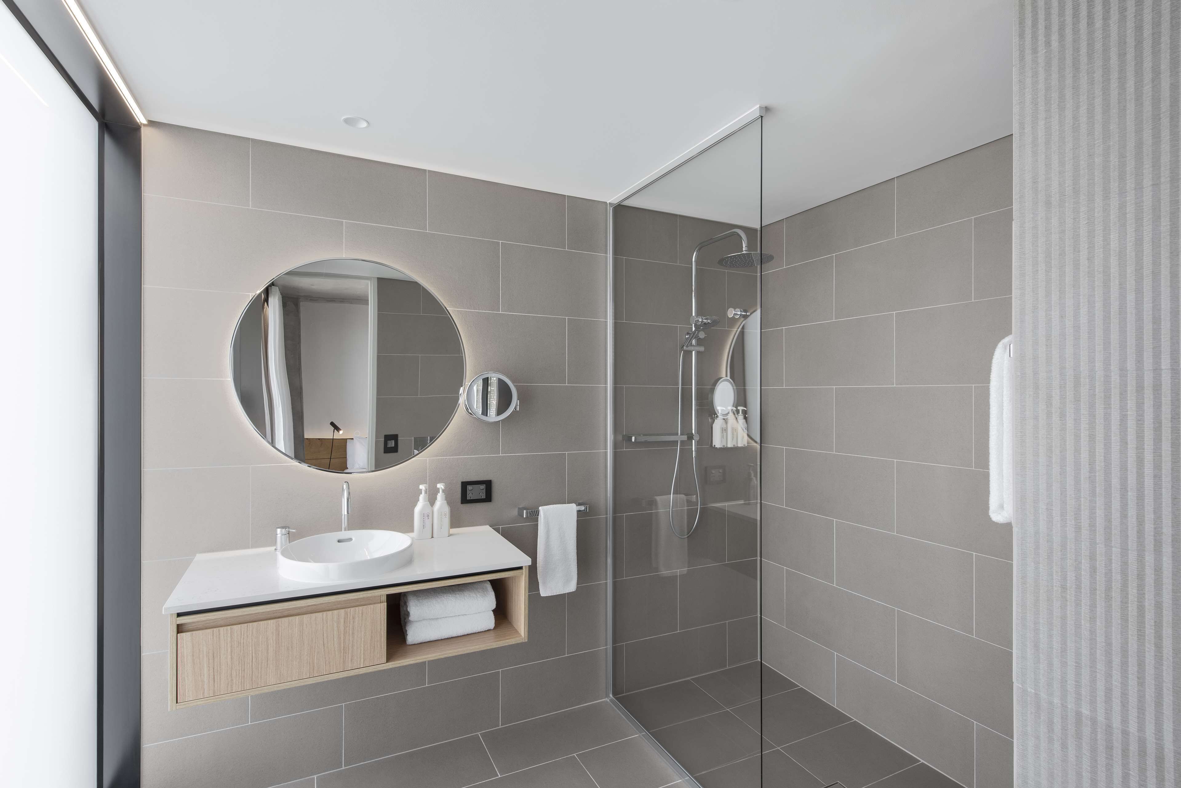 A modern bathroom with a glass-enclosed shower, a round mirror, and a white vanity with a sink and towel rack.