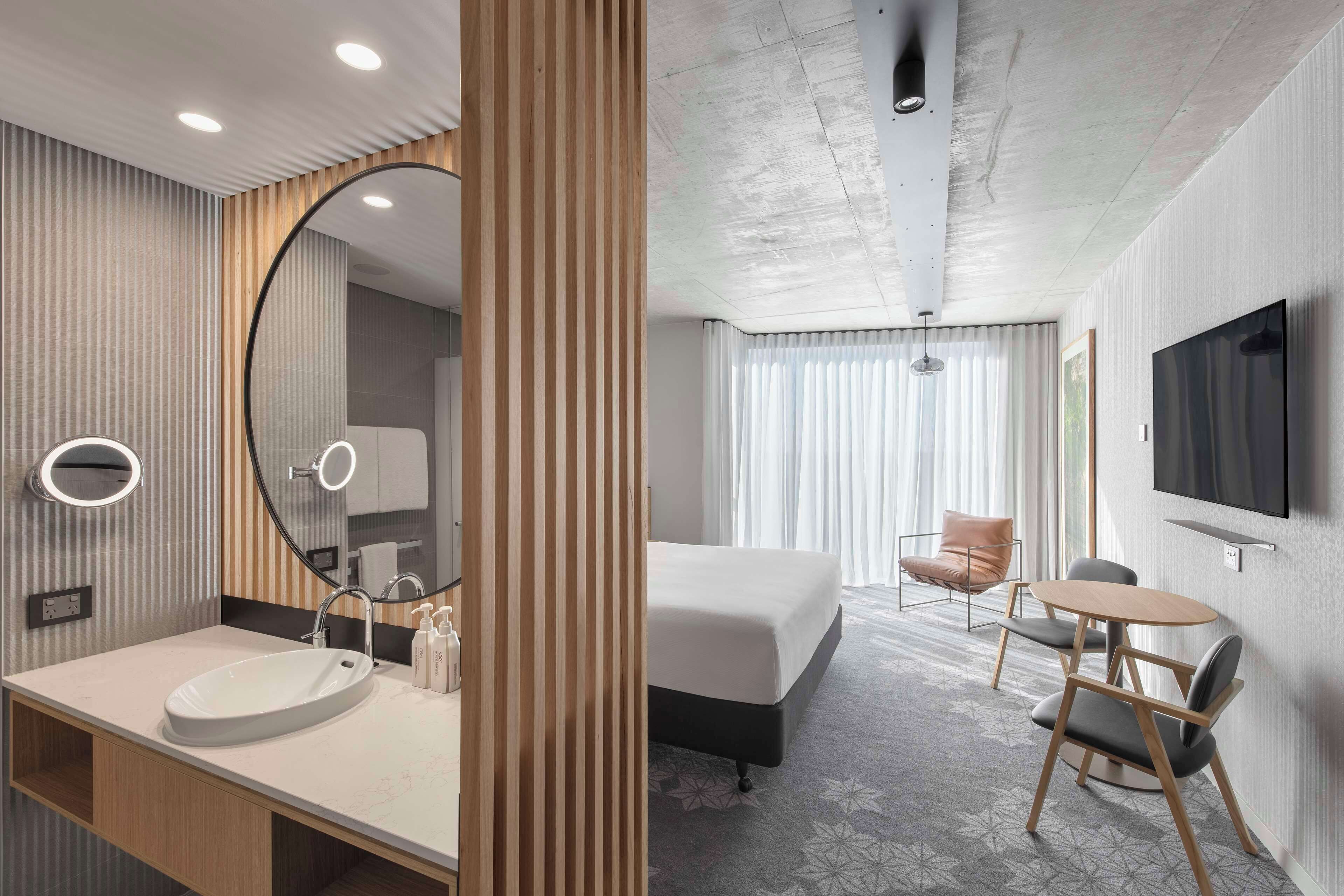 A hotel room with a large bed, a round table, two chairs, a TV mounted on the wall, and a bathroom with a large mirror and sink.