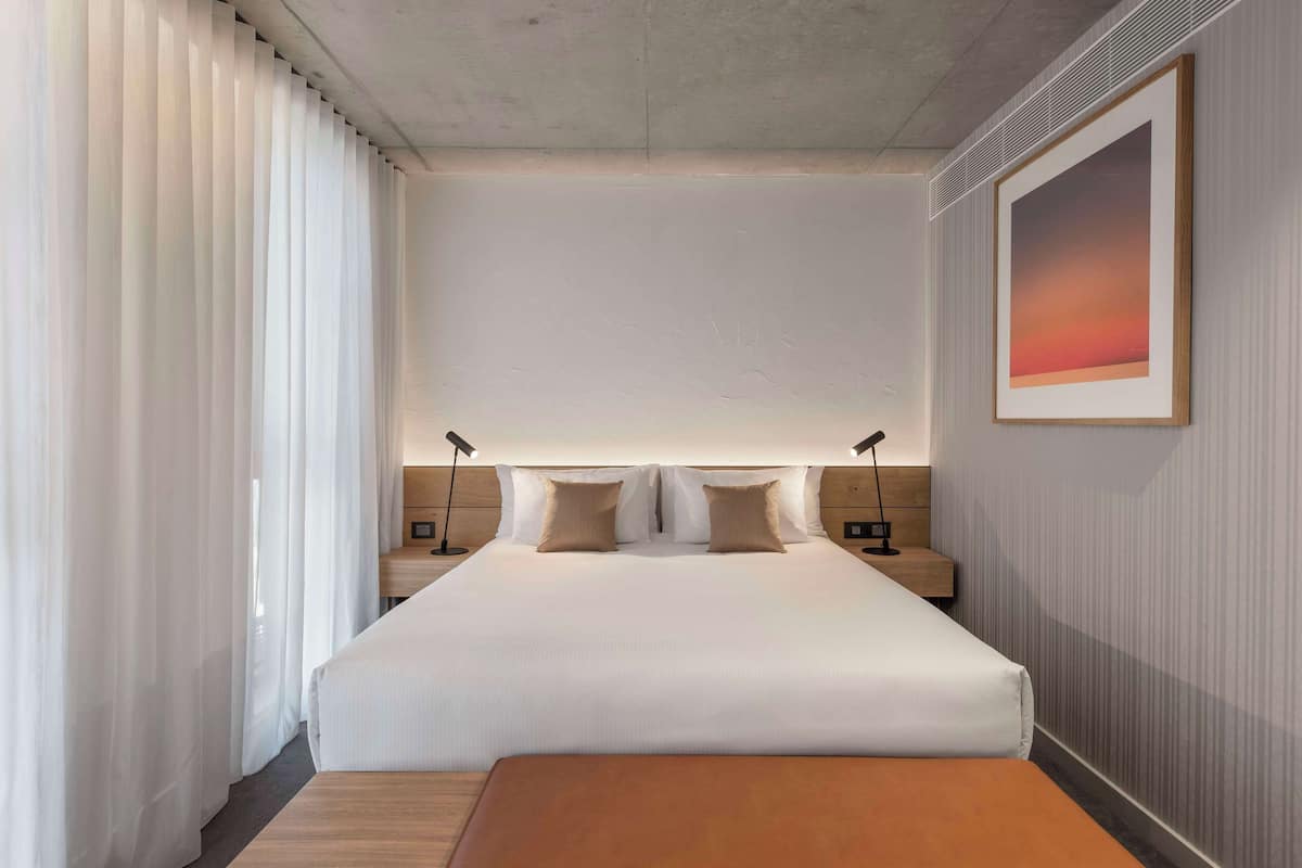 Habitación de hotel moderna con una cama grande, dos mesitas de noche con lámparas, un banco de madera y un cuadro grande en la pared.