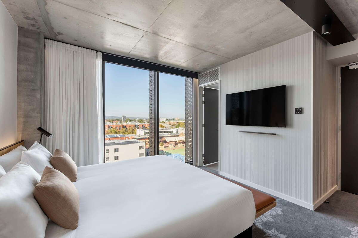 A modern hotel room with a large bed, a flat-screen TV mounted on the wall, and a city view through the window.