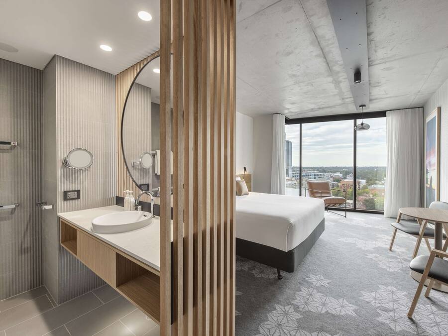 Una habitación de hotel moderna con una cama grande, un baño con lavamanos y espejo, y vistas a la ciudad a través de amplias ventanas.