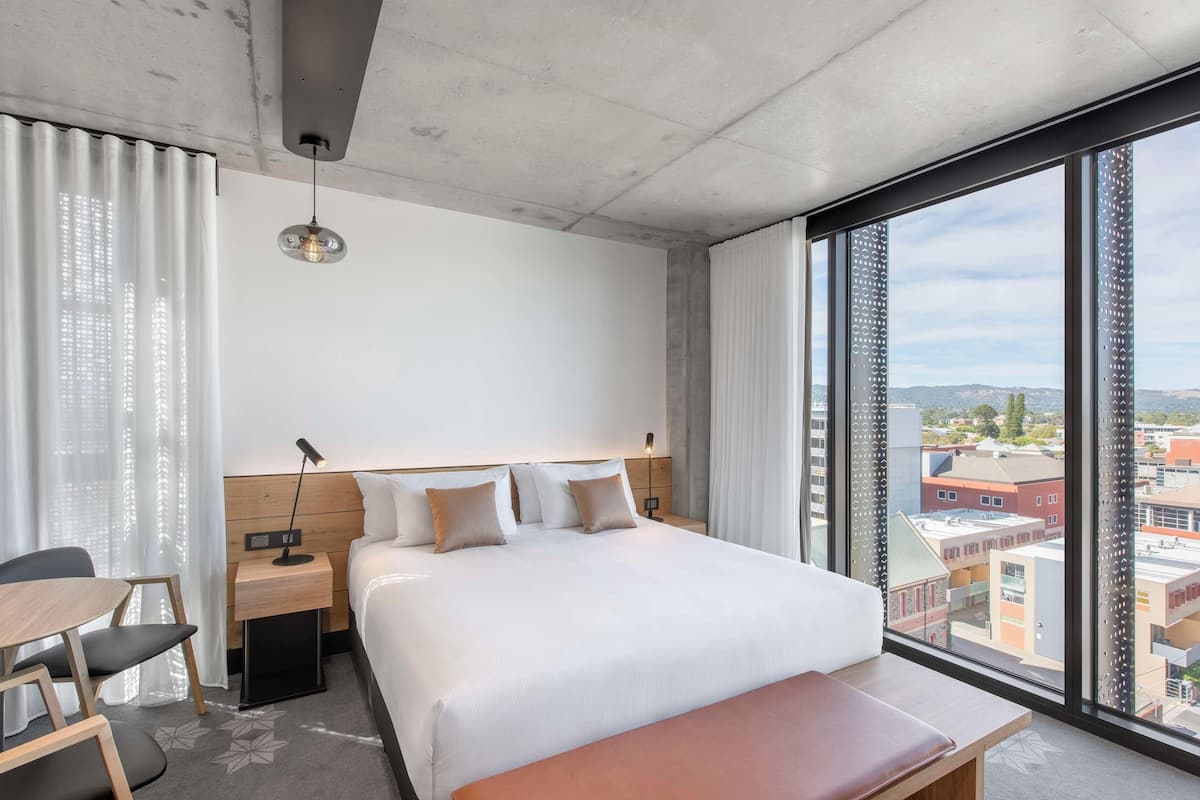 Habitación de hotel con una cama grande, un escritorio, una silla, una mesita de noche, una lámpara y vistas a la ciudad a través de amplias ventanas.