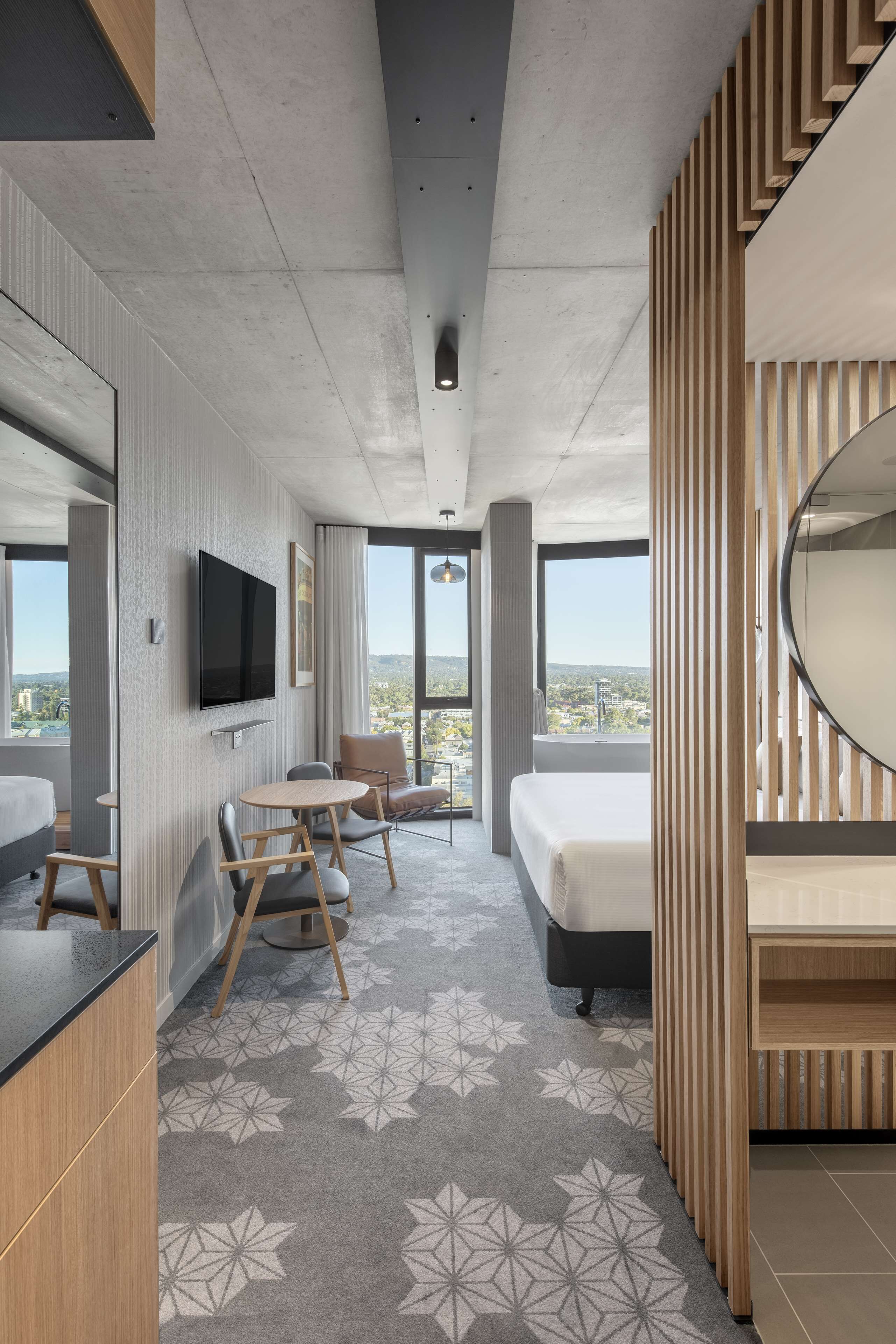 A modern hotel room with a large bed, a desk with chairs, a flat-screen TV mounted on the wall, and a large window offering a city view.