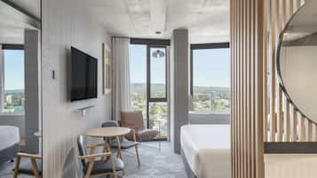 A modern hotel room with a large bed, a desk with chairs, a flat-screen TV mounted on the wall, and a large window offering a city view.