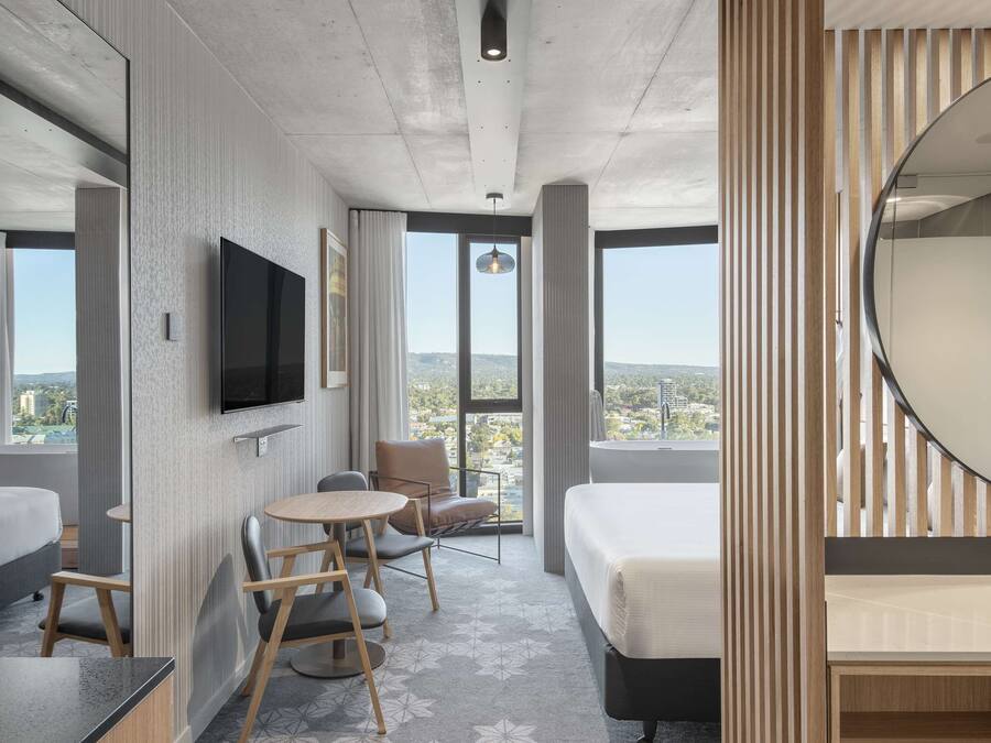 Ein modernes Hotelzimmer mit einem großen Bett, einem Schreibtisch mit Stühlen, einem flachbildfernseher an der Wand und einem großen Fenster mit Blick auf die Stadt.