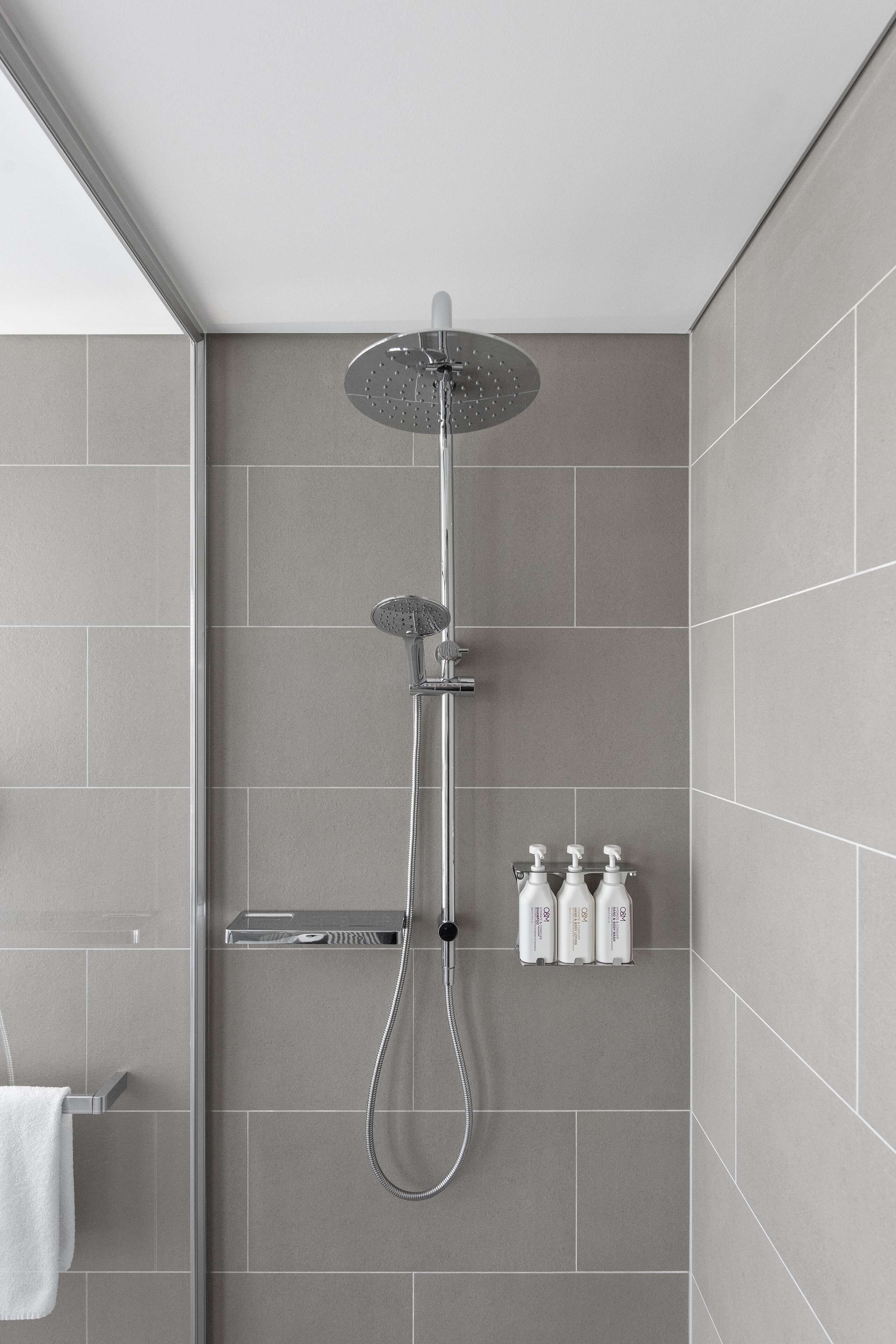 Ducha moderna con cabezal de ducha de lluvia, cabezal de ducha manual y un accesorio de ducha manual. Hay tres botellas de champú y acondicionador sobre una repisa.