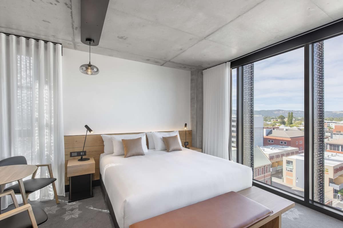 Una habitación de hotel moderna con una cama grande, un escritorio y vistas de la ciudad a través de amplias ventanas.