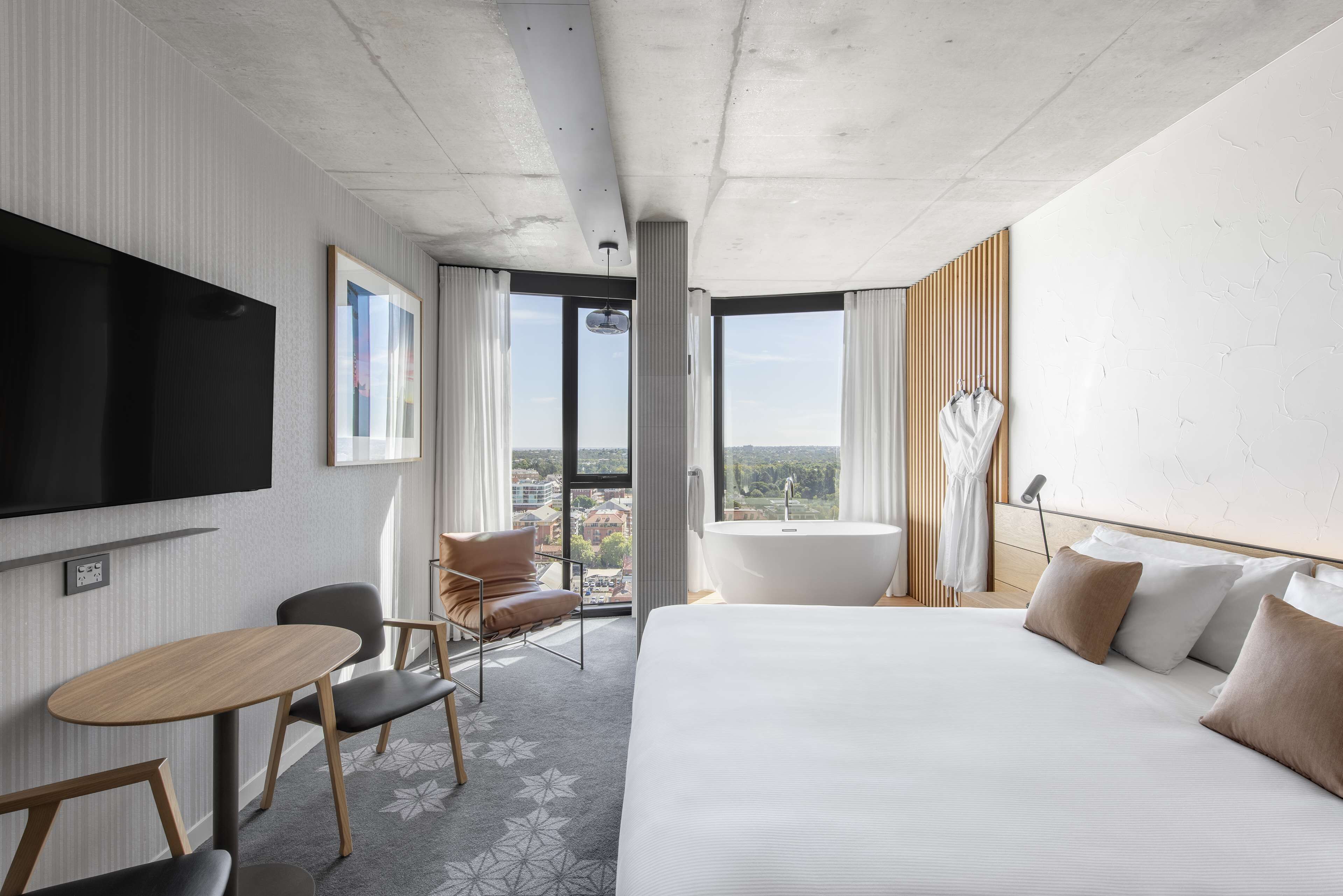 Una habitación de hotel moderna con una cama grande, un televisor de pantalla plana, un área de descanso con una mesa y sillas, un ventanal con vista a la ciudad y una bañera.