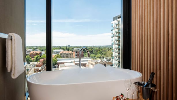A modern bathroom with a freestanding bathtub, wooden flooring, a wooden side table with a wine glass and bottle, and a large window offering a city view. - Vibe Hotel Adelaide (Adelaide)