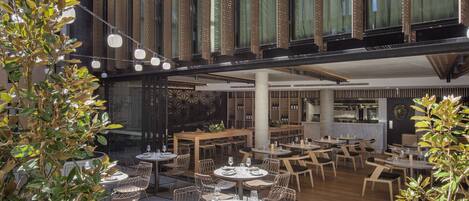 Un área de comedor al aire libre con mesas y sillas, rodeada de grandes macetas y un edificio moderno con ventanales grandes y una fachada decorativa.