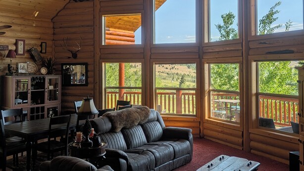 A log cabin interior with a high ceiling, large windows, a fireplace, a dining area, and a cozy seating area.