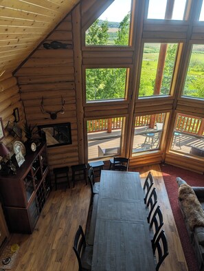 A log cabin interior with a dining table, chairs, a fireplace, and large windows offering a view of a green landscape.