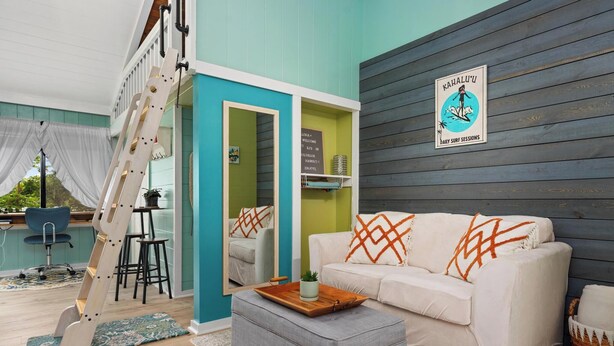 A living room with a sofa, ottoman, ladder, and a sign for daily surf sessions. - Adorable Cottage w/ Jungle & Mountain Vibes (Holualoa)