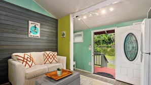 A room with a sofa, a small table, a door leading outside, and a wall-mounted sign. - Adorable Cottage w/ Jungle & Mountain Vibes (Holualoa)