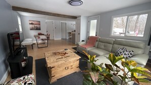 Una sala moderna con una mesa de centro de madera, un sofá gris con cojines estampados, un comedor con mesa y sillas, y un ventanal con vistas a árboles.
