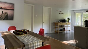 Una cocina moderna con una mesa de madera, sillas rojas y un cuenco de fruta. Hay un cuadro en la pared y vistas al exterior a través de un ventanal.