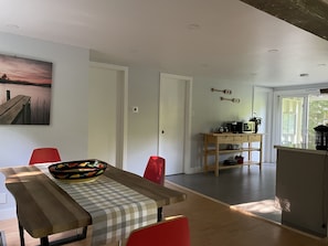 A modern kitchen with a wooden table, red chairs, and a fruit bowl. There is a painting on the wall and a view of the outdoors through a glass door.