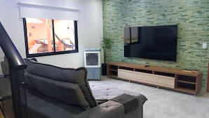 A living room with a grey sofa, a flat-screen TV mounted on a green brick wall, a wooden entertainment unit, and a window with a view of outdoor furniture. - Beautiful Townhouse with 3 suites, Pool and Garage for 4 Cars (Florianópolis)