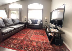 A living room with a large flat-screen TV, two leather sofas, a red patterned rug, and a small side table.