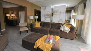A modern living room with a brown sofa, a matching ottoman, and a coffee table with a decorative item. There is a large mirror and a lamp on a side table. - 224 Calle Florista, Laguna De LaPaz (La Quinta)