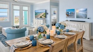 A dining area with a wooden table set for six, a TV mounted on the wall, and a view of the ocean through large windows. - Pointe West Family Beach + 2 Resort Pools (Galveston)