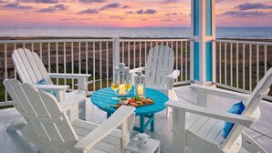 A deck with white Adirondack chairs, a blue table, and a view of the ocean at sunset. - Pointe West Family Beach + 2 Resort Pools (Galveston)