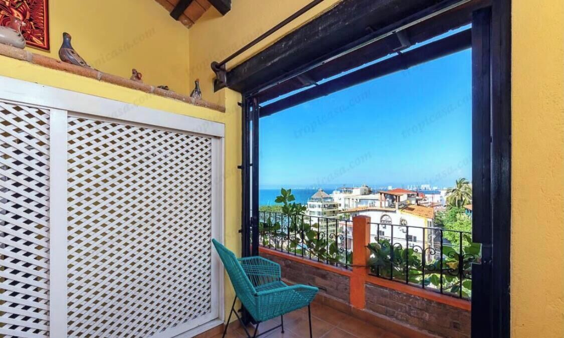 Un balcón con vista a la ciudad, un separador de celosía blanca y una silla de mimbre verde.
