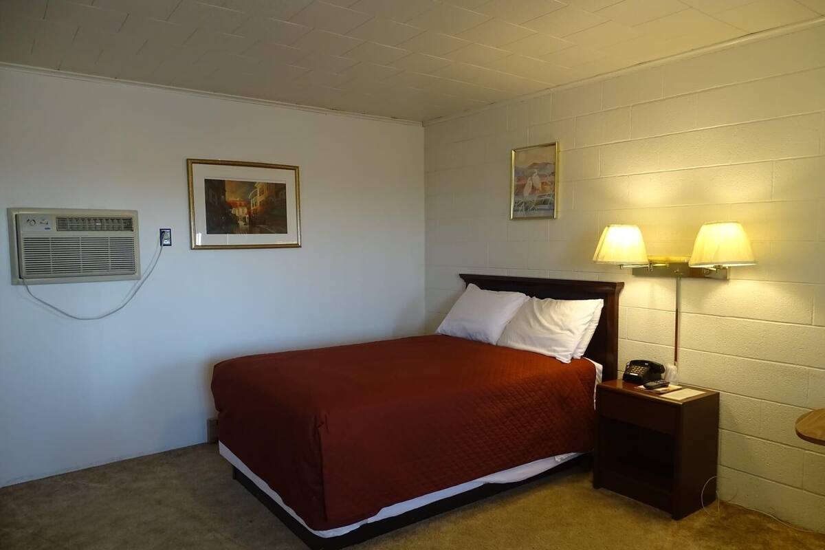 Uma cama de solteiro com colcha vermelha em quarto com paredes brancas, ventilador de teto e duas luminárias fixadas na parede.