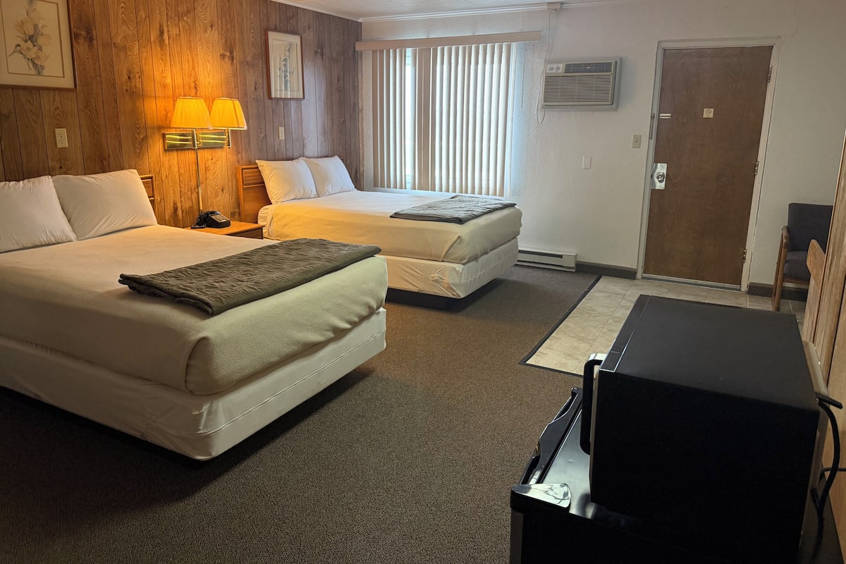 Quarto de hotel com duas camas, uma escrivaninha, uma luminária, uma TV e uma janela com persianas.