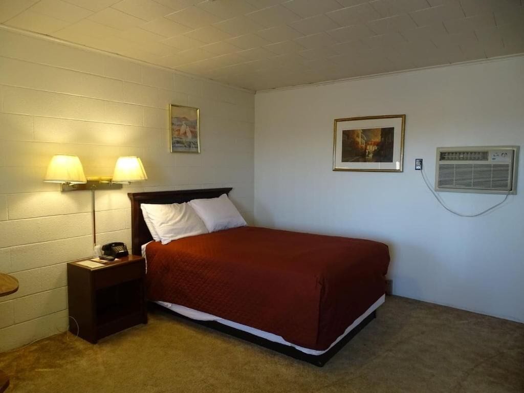 Uma cama de solteiro com um cobertor vermelho, duas luminárias na parede, um quadro emoldurado e um aparelho de ar condicionado.