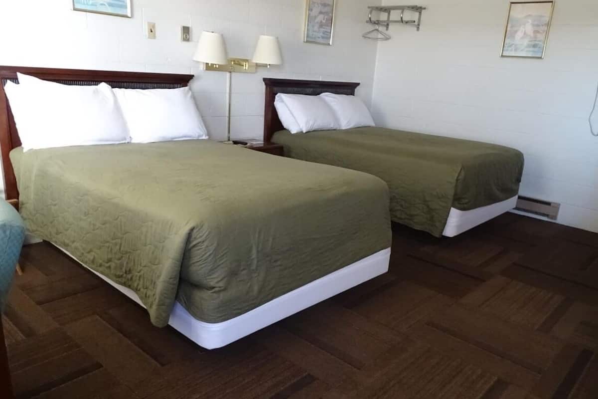 Quarto de hotel com duas camas de solteiro, mesa de cabeceira, abajur e prateleira na parede.