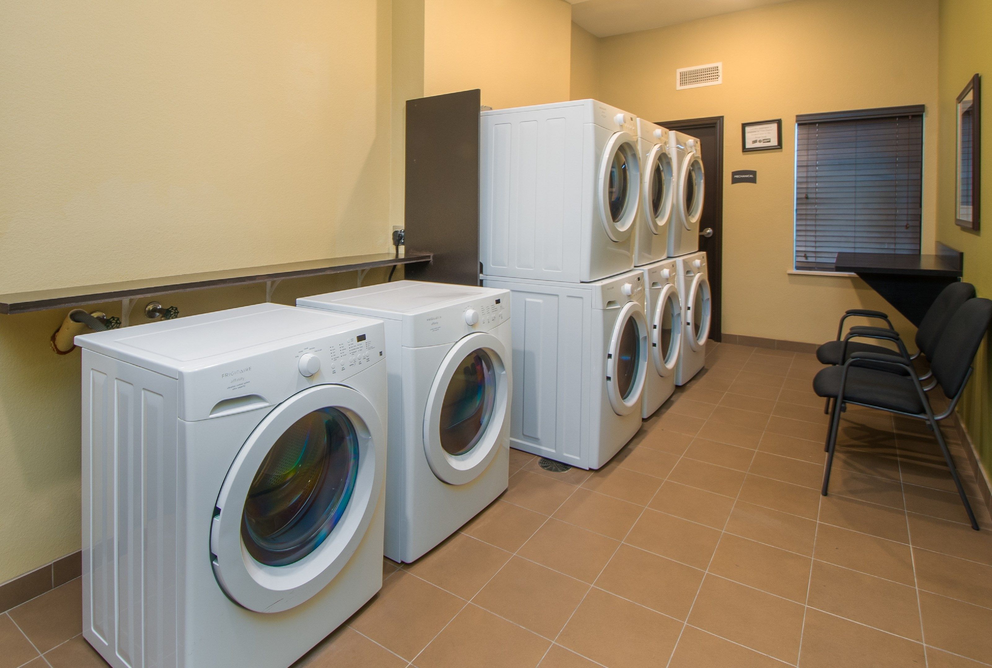 A laundry room with multiple washing machines and dryers, a chair, and a window with blinds.